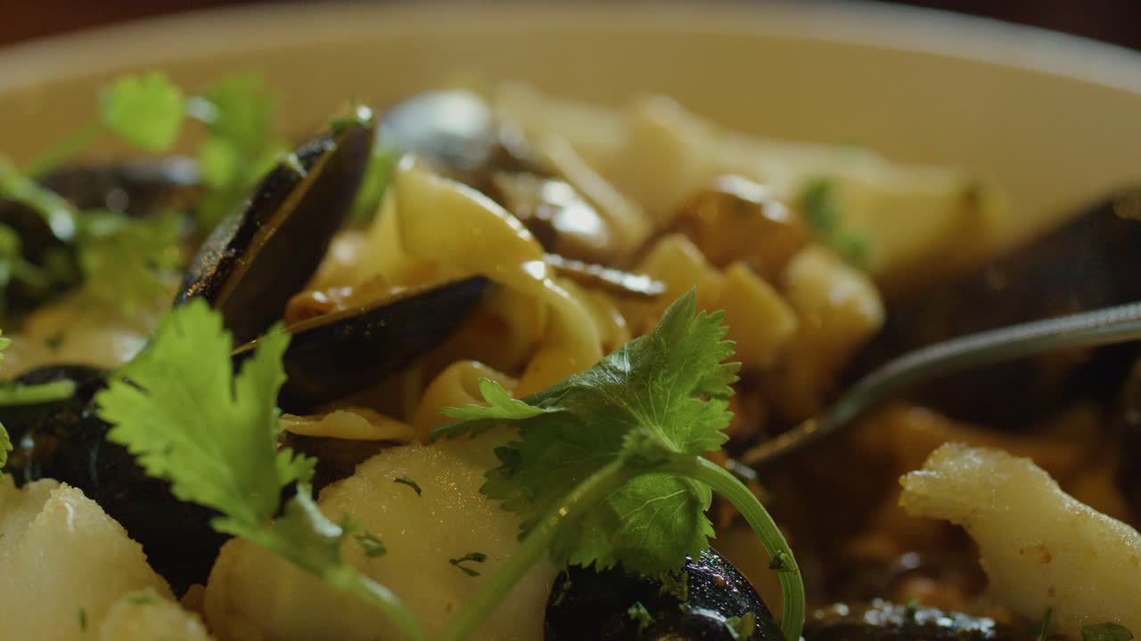 A hand uses a spoon to scoop steaming seafood chowder with fish, squid, mussels, and herbs in warm, natural lighting. Macro perspective highlights textures