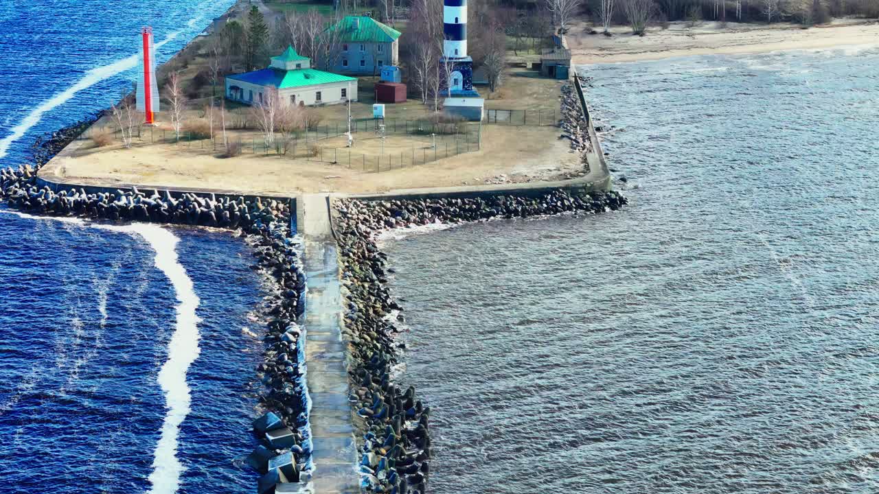 A black and white lighthouse stands beside simple houses on a long stone jetty splitting vivid blue Baltic Sea from muted estuary waters along Latvia's rugged coastline.