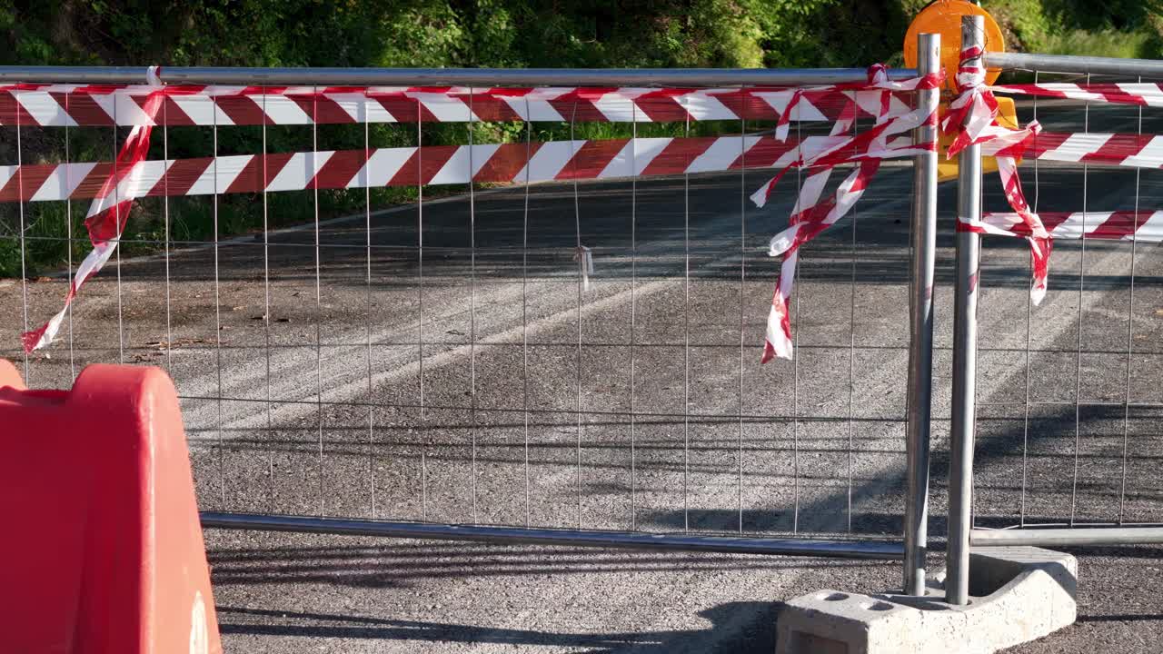 A metal barrier with red and white caution tape blocks a section of asphalt road, indicating temporary closure or construction. The scene is lit by direct sunlight.
