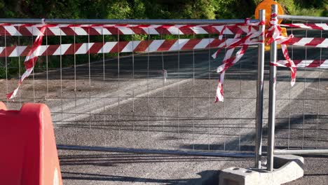 a metal barrier with red and white caution tape blocks a section of asphalt road, indicating temporary closure or construction. the scene is lit by direct sunlight.