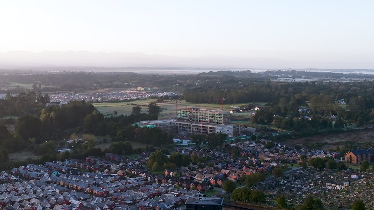 Luftaufnahmen einer Drohne bei Sonnenaufgang, die die Baustelle des zukünftigen Krankenhauses in Puerto Varas, Chile, mit Kränen und Morgenlicht zeigen.
