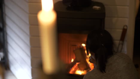 Woman-stares-thoughtfully-into-flames-of-home-fireplace,-candle-flickers-in-foreground