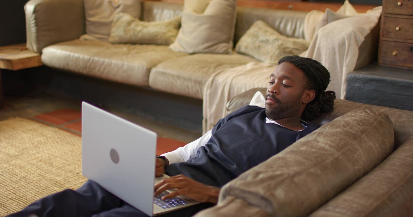 Cam panning African-American man lounging sofa arm typing laptop working online at home, copy space