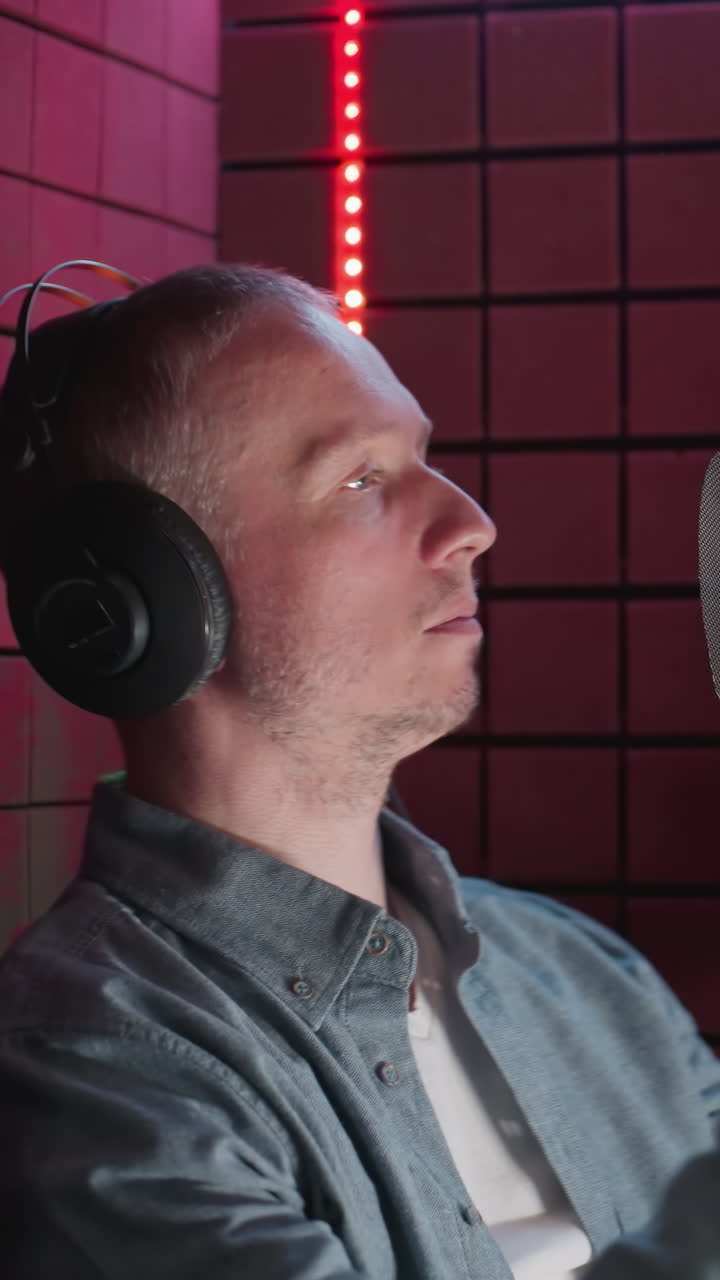 un hombre con una camisa azul y una camisa interior blanca está en una habitación a prueba de sonido roja, usando auriculares, y moviendo su cuerpo mientras canta en un micrófono