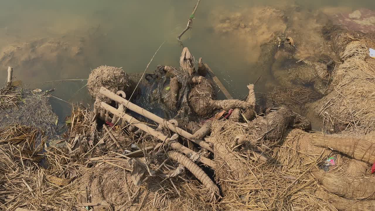 Weathered idol remnants made of straw and bamboo lie tangled along a muddy riverbank, partially submerged in still, murky water