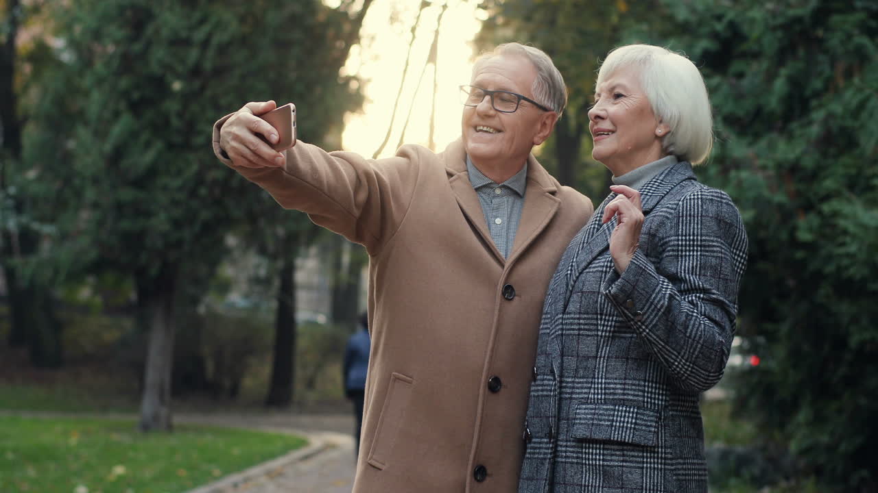 pareja de ancianos posando y sonriendo a la cámara mientras toma una foto selfie con un teléfono inteligente en el parque al atardecer