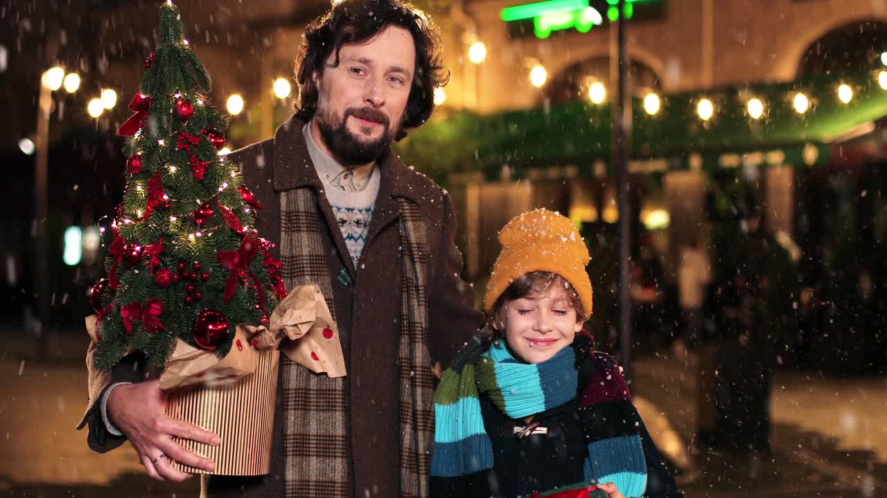 retrato de padre y su hijo pequeño sosteniendo un regalo y un árbol de navidad, y mirando la cámara en la calle mientras nieva en navidad
