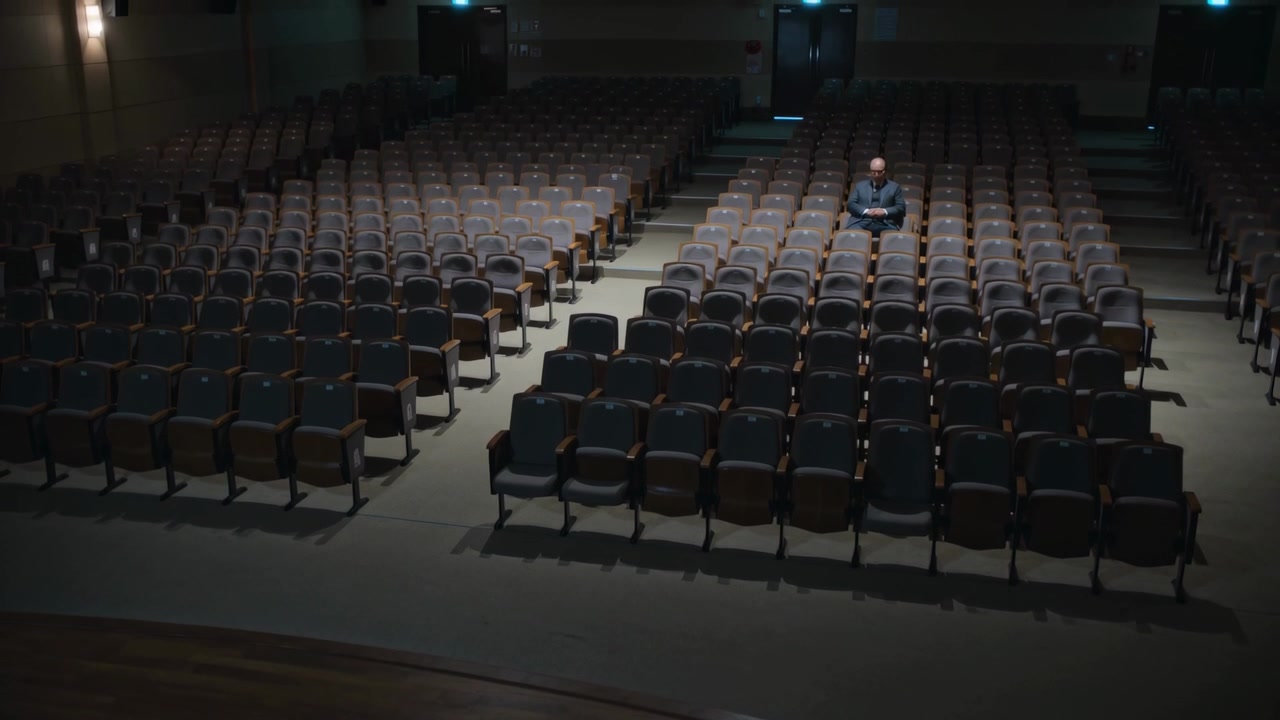 Uomo seduto in giacca scura che guarda il palco in una grande sala, in attesa dell'inizio della lezione tra sedili vuoti