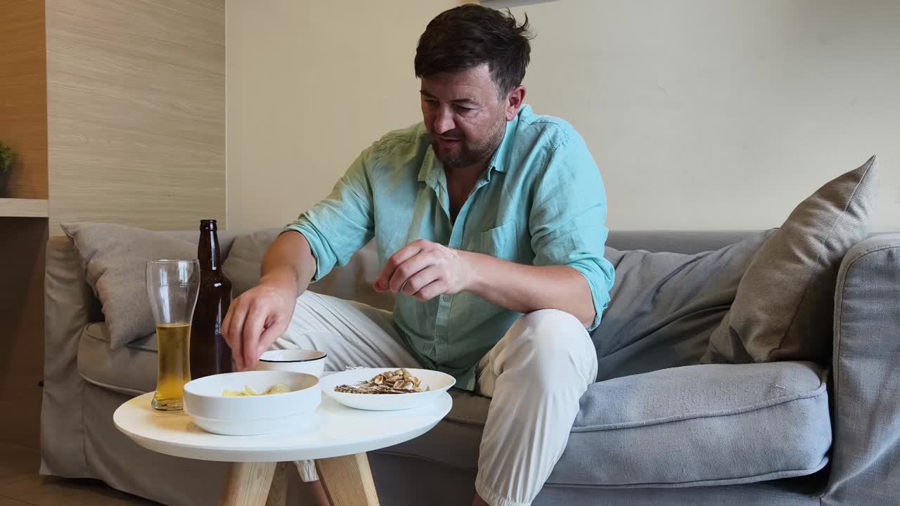 A man relaxes on a couch, eating snacks and drinking beer