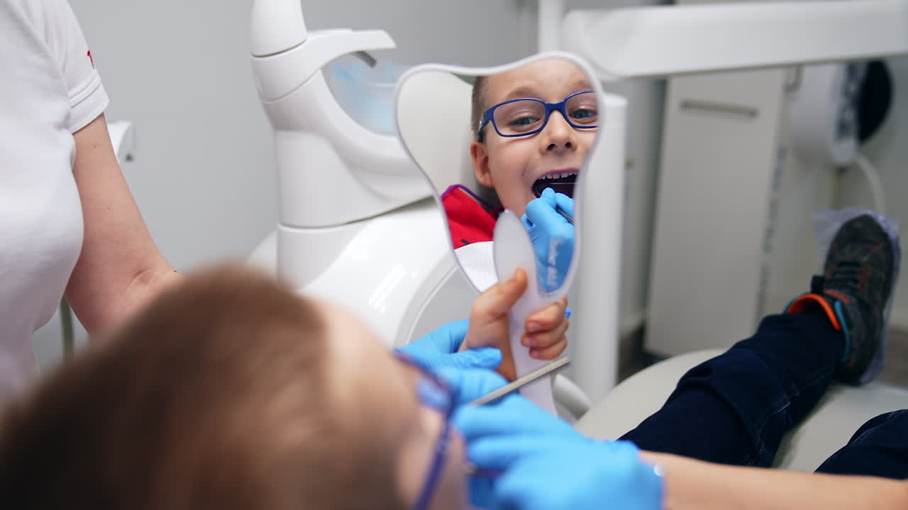 Smiley little boy looking at himself in the mirror. Dentist showing the result of her work with the help of metal tools.