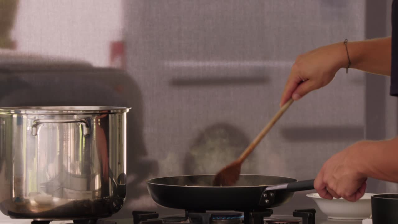 A close-up shot of hands stirring a pan on a stove in a kitchen, with a stainless steel pot nearby, capturing the cooking process in a professional setting.