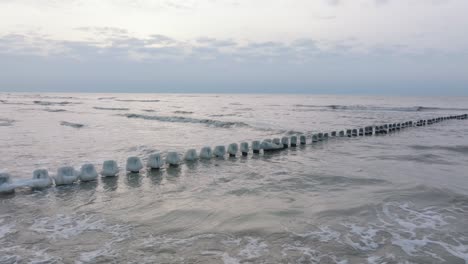 Aerial-establishing-view-of-an-old-wooden-pier-at-the-Baltic-sea-coastline,-overcast-winter-day,-white-sand-beach-covered-in-snow,-ice-on-wood-poles,-calm-seashore,-wide-drone-shot-moving-forward-low