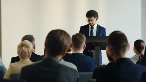 caucasian businessman wearing glasses and formal clothes speaking at a conference in front of many people