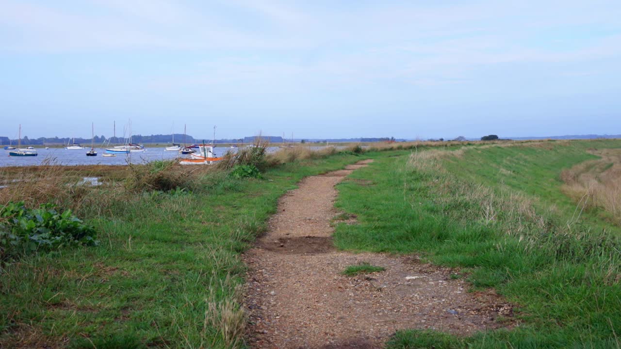 Tilt up to show river wall defence. Dirt path on man made grassy hill to defend lowland farms in England. Water and boats on left side. Blue sky with clouds.