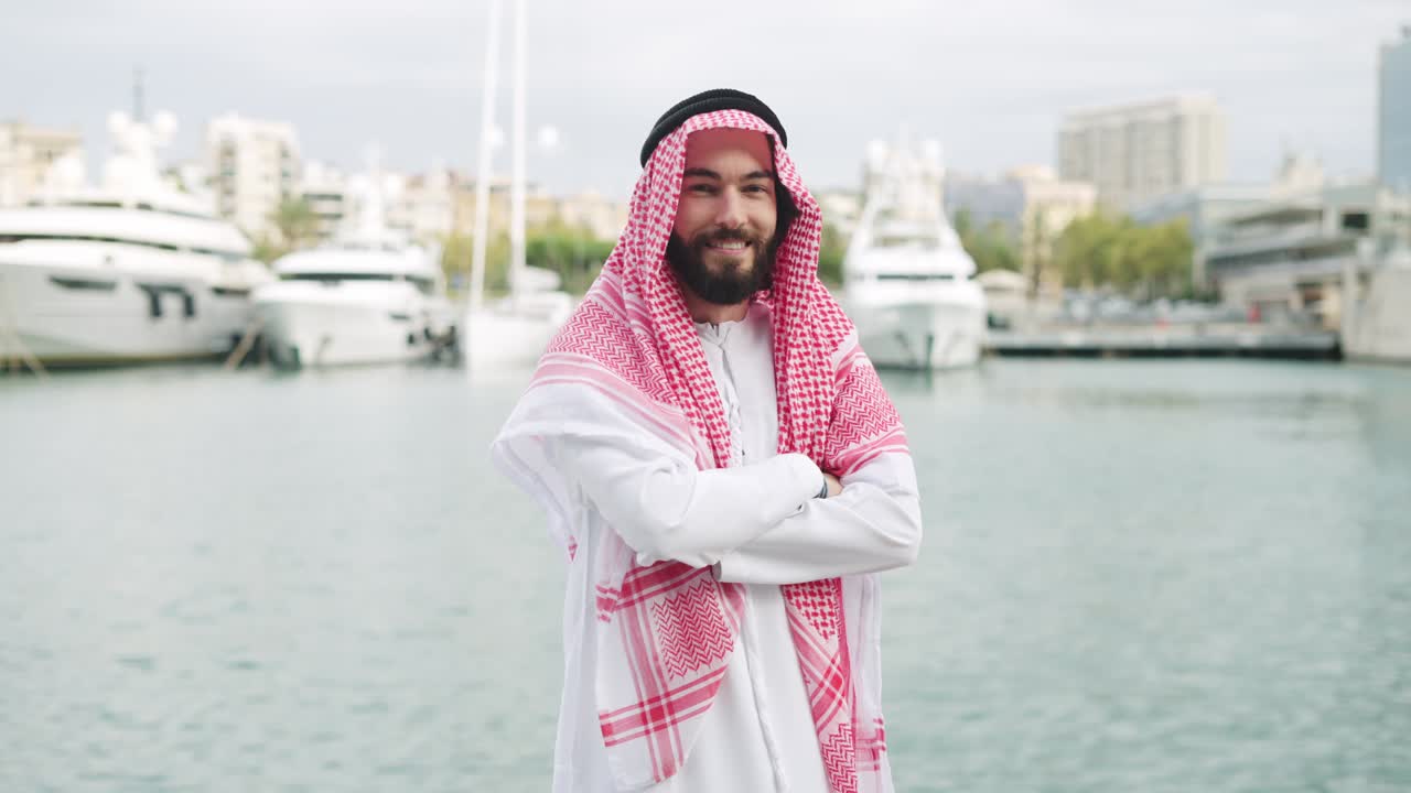feliz hombre saudí sonriente posando al aire libre con ropa tradicional con los brazos cruzados en el puerto