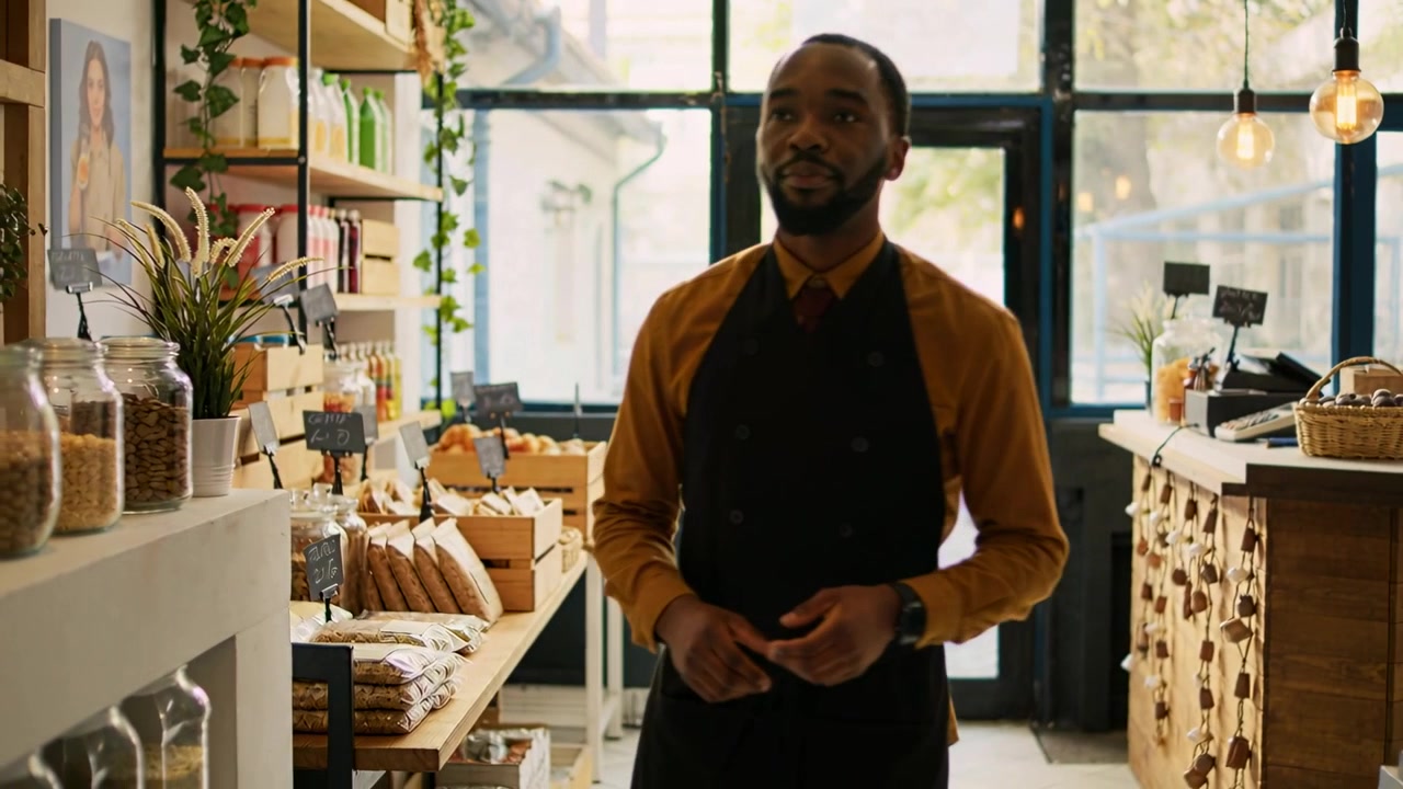 Un employé en tablier foncé rangeant l'étagère de gauche dans un magasin, garnissant des bocaux et guettant les clients à la porte.