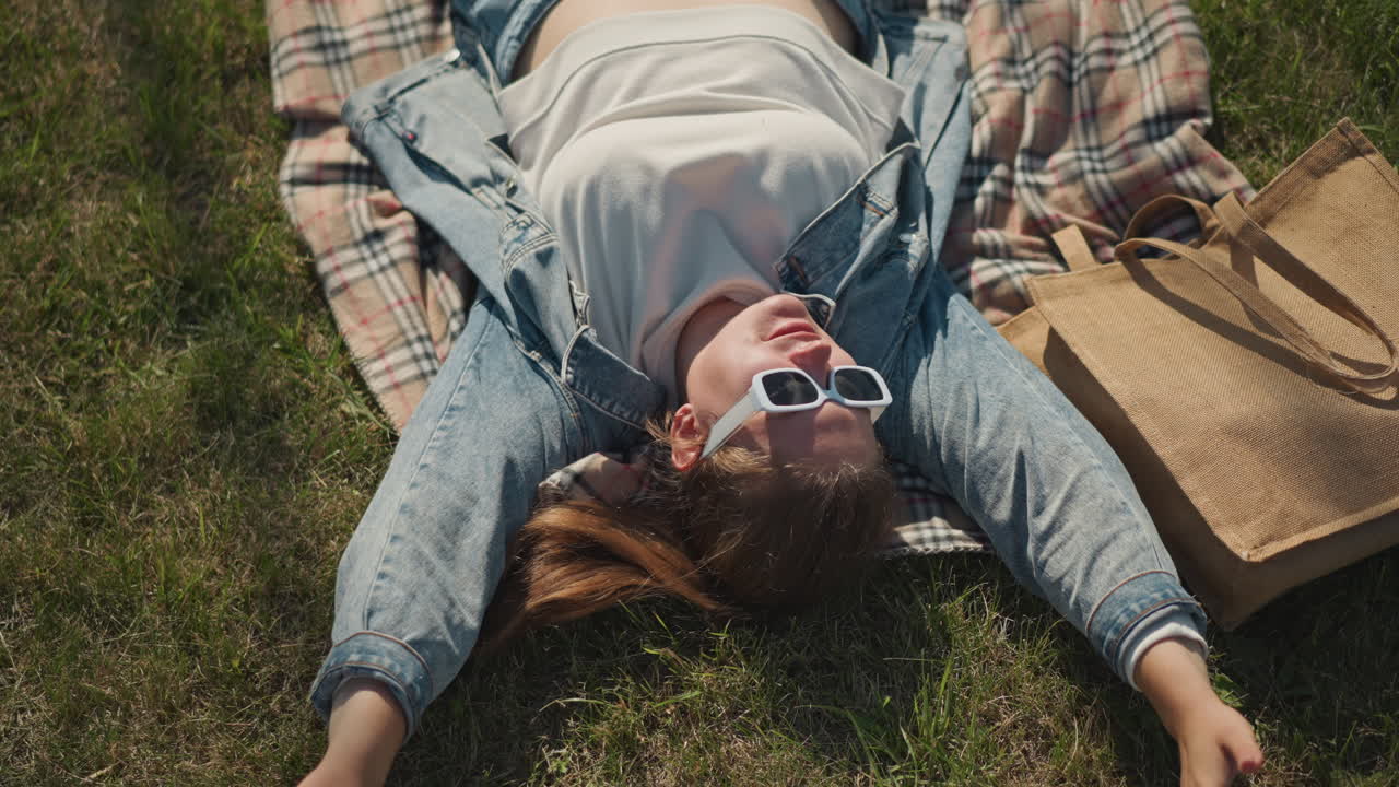 White woman lying on grass blanket, wearing sunglasses and denim jacket, straw tote beside her, sunlight dappling plaid blanket, overhead view of relaxed student daydreaming during picnic break