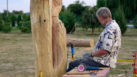 grey-haired bearded man sits on the bench near a piece of wood outdoors. artist draws lines on the timber with a chalk.