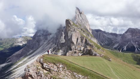 Vista-Aérea-De-Un-Excursionista-En-La-Cima-De-La-Montaña-Verde-De-La-Caminata-De-La-Cresta-De-Seceda-Viendo-Las-Montañas-Dolomitas-Durante-El-Día-Nublado,-Italia