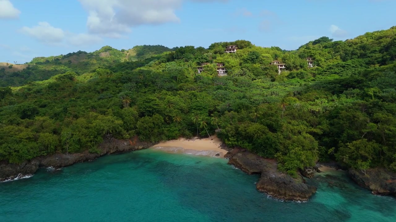 Spiaggia appartata a forma di cala che si curva tra scogliere rocciose a Playa Bremen a Las Galeras, Samaná, Repubblica Dominicana, con ville collinari che fanno capolino attraverso la fitta chioma verde sopra la riva. Vista aerea con drone