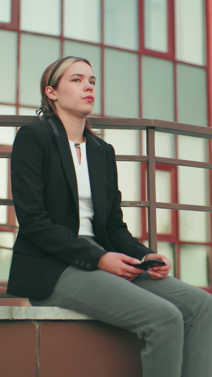 Lady holding phone seated on fence in urban area with striking modern building behind, metal railings around, calm expression, natural light, professional attire, reflective mood in outdoor setting