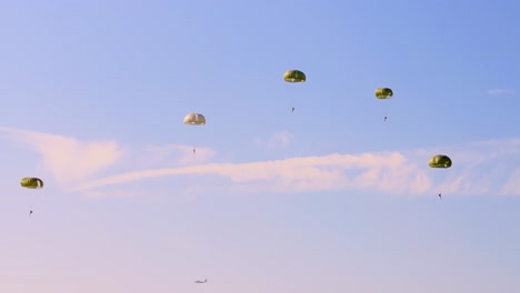 four parachutists drift gracefully under green canopies, descending from an airborne operation into a clear blue sky with light cloud streaks