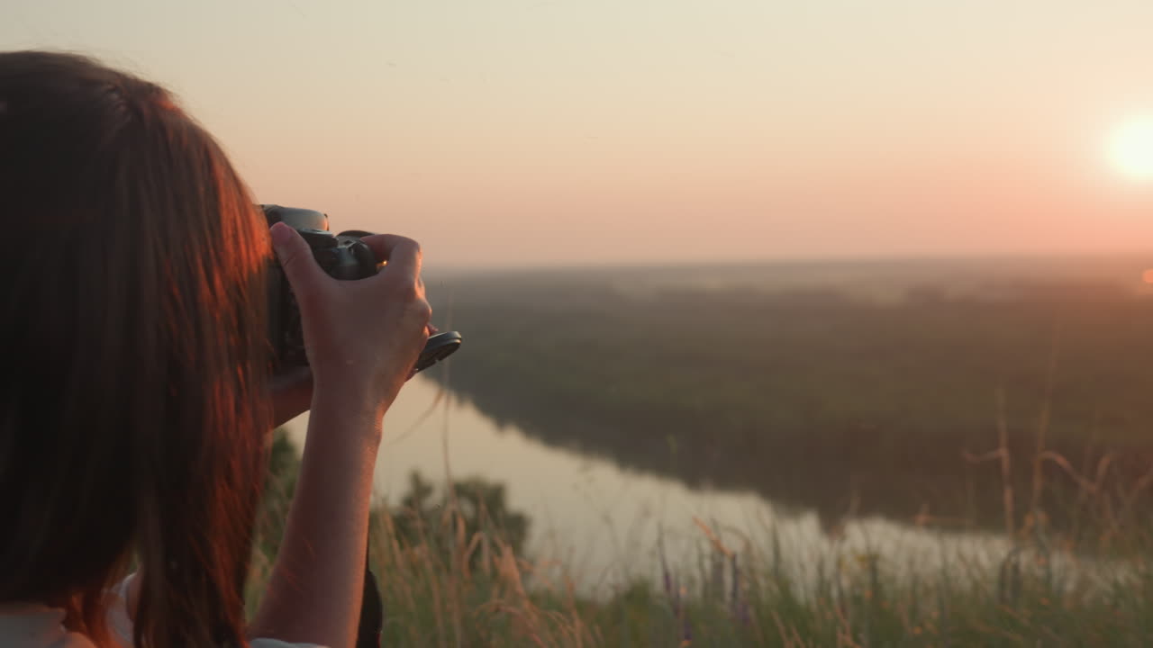 Rear view of photographer capturing scenic sunset horizon over blurred river and lush green banks, holding DSLR camera with long lens steady while framing warm golden sky and tranquil flowing water
