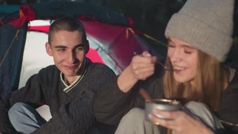 young man watching woman eating food from can while sitting outdoors near camping tent during evening relaxed atmosphere nature adventure couple sharing meal enjoying outdoor lifestyle