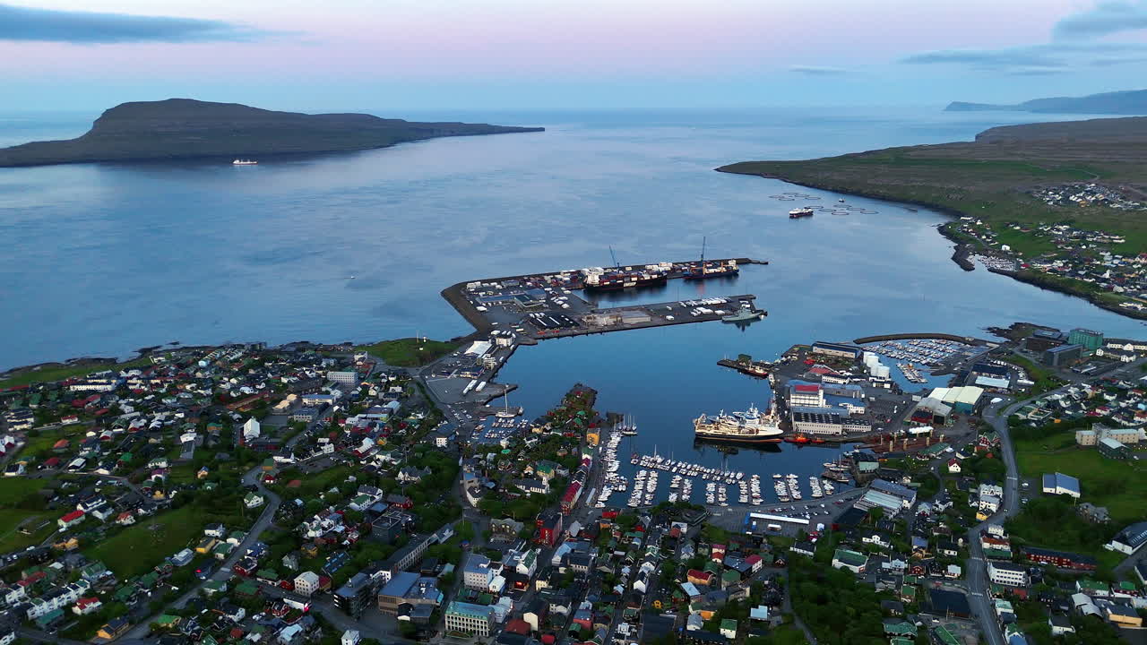 Cinematic aerial view of colorful village houses along a fjord in the Faroe Islands, surrounded by dramatic green mountains, black sand beach, and misty coastal scenery