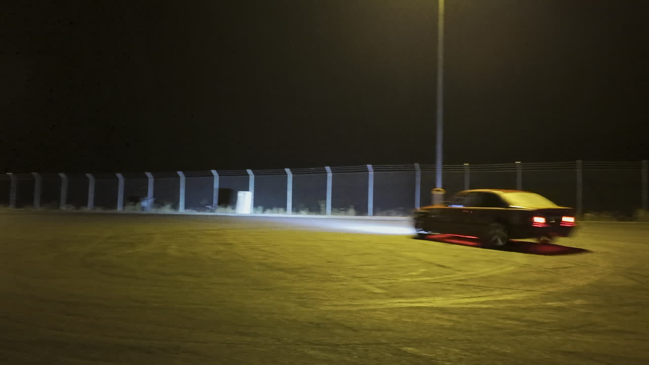 A fast sports car performing a drift in an open lot illuminated by bright streetlights on a rainy night
