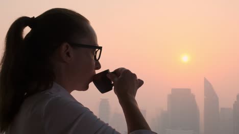 dreaming woman drinks tasty coffee while looking out window at evening city at sunset