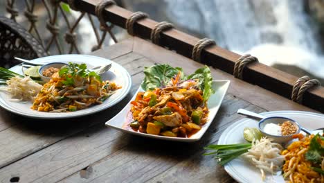 traditional thai food in outdoor cafe with nature waterfall background. fried noodle with prawn pad thai and chicken cashew nut on wooden table in restaurant. asian style food lunch.