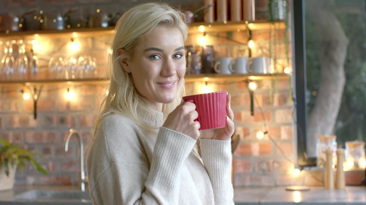 Entrando a la cocina, mujer sostiene una taza roja, sorbe café, mira por la ventana y sonríe a la cámara.