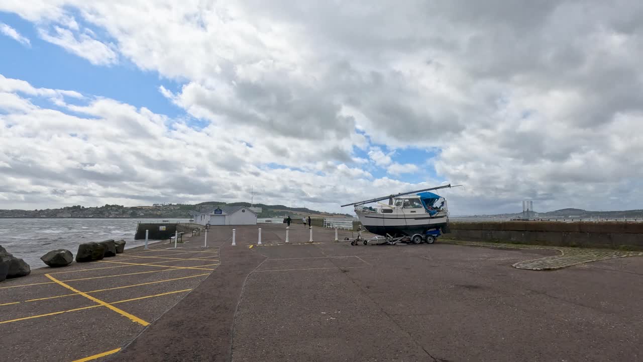 A motor sailer boat is moved on a trailer across an empty coastal parking lot under partly cloudy daylight, with steady wide-angle camera framing
