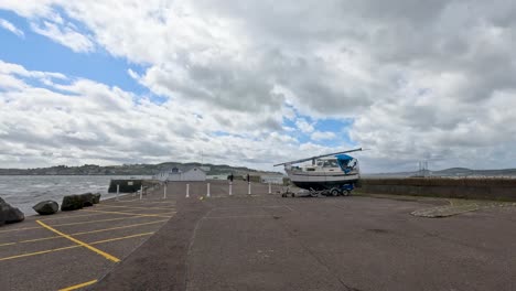 a motor sailer boat is moved on a trailer across an empty coastal parking lot under partly cloudy daylight, with steady wide-angle camera framing