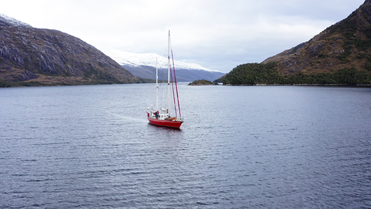 Sail boat floats in calm waters of Beagle Channel surrounded by distant hills and clouds, aerial establishing medium