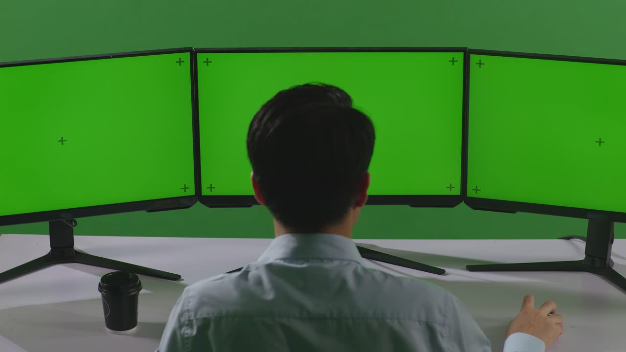 행복한 아시아 남자가 축하하고 춤을 추고 있는 뒷면으로 녹색 스크린 스튜디오에서 여러 컴퓨터 모니터를 모방하는 작업을 하고 있습니다.