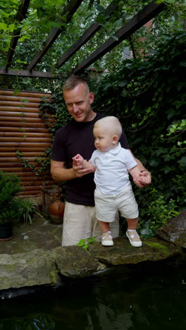 father holds his little baby standing at the fountain. cute infant kid looking at water flowing. beautiful greenery at backdrop. vertical video.