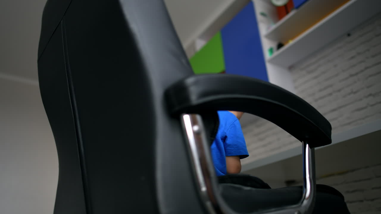 Cute toddler boy sits in a office chair with phone in hands. Barefoot kid is rotated in the chair. Low angle view.