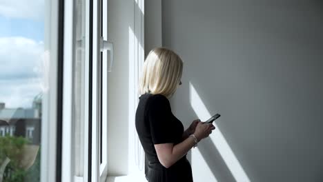 mujeres rubias anónimas inclinadas junto a la ventana de la sala de reuniones usando su teléfono inteligente