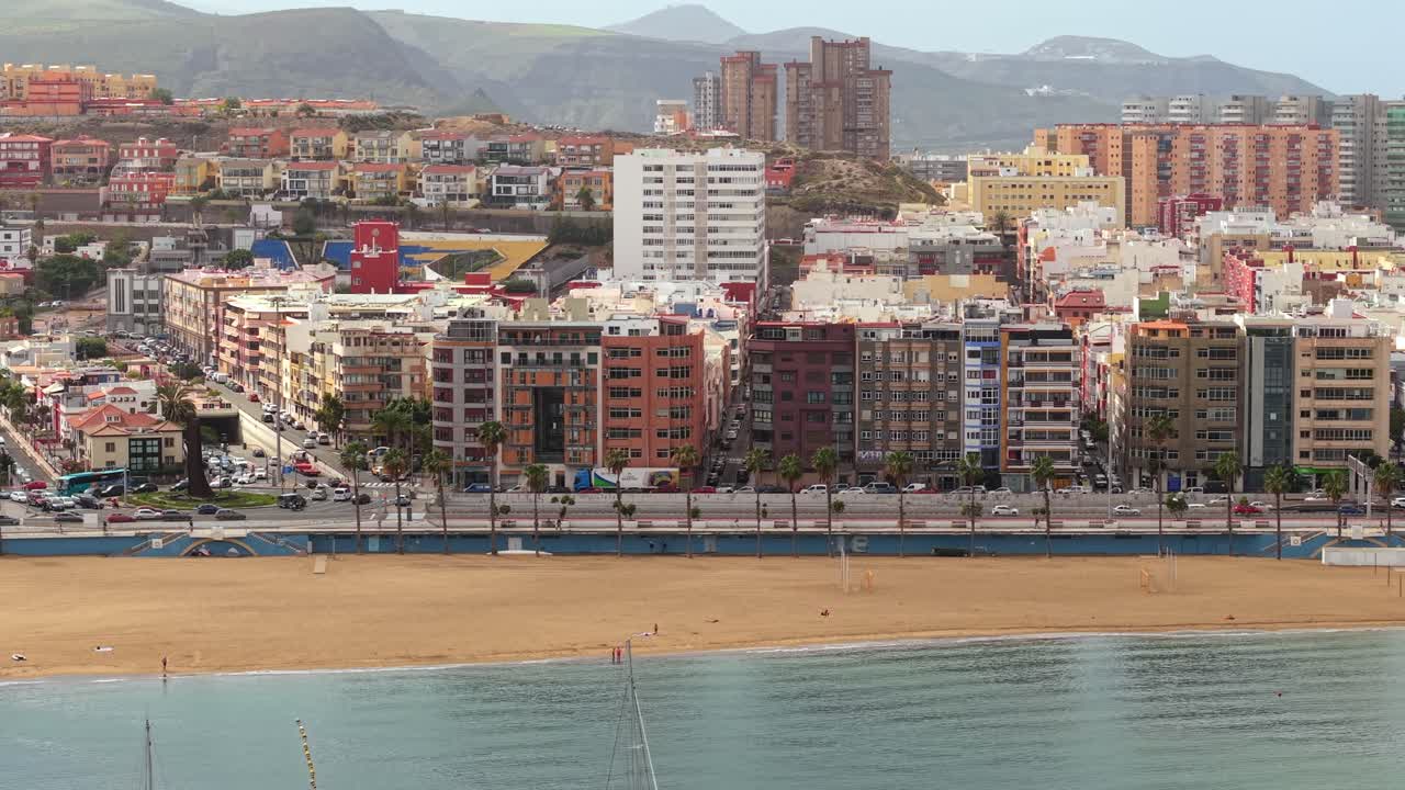 Panoramisk luftperspektiv af bylandskabet i Las Palmas, der viser den urbane Alcaravaneras-strand, havnefrontsbygninger og bjerge.