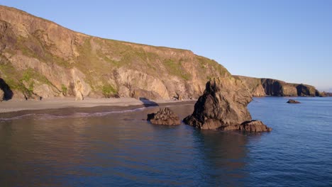 drone circling sea stack with dramatic cliffs and shingle beach at high tide at golden hour copper coast waterford ireland