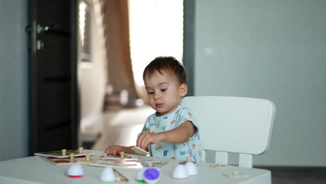 cute little toddler plays with toy sitting at desk. lovely boy collects puzzles and claps hands happily.