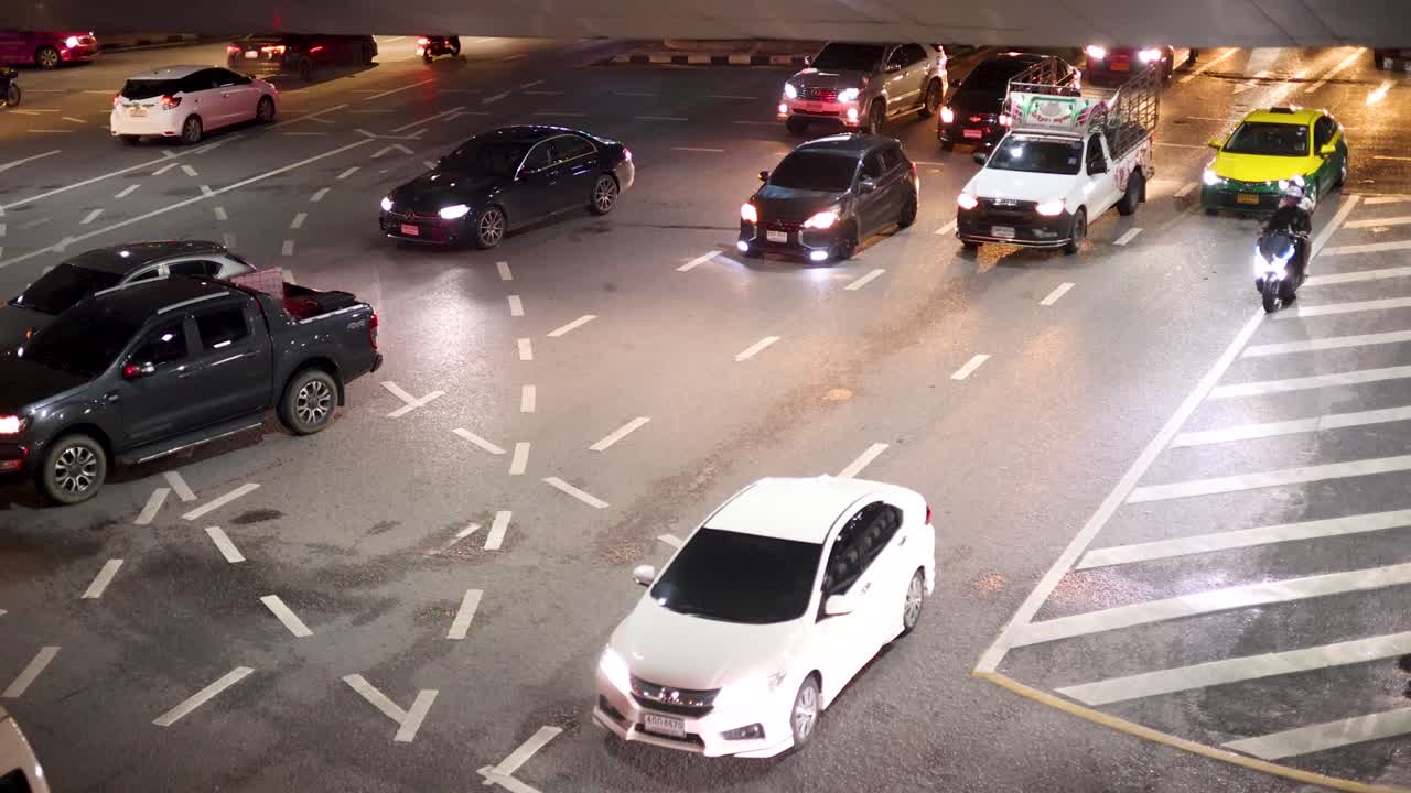 Multiple vehicles and motorcycles navigate a brightly lit Bangkok intersection at night, overhead view