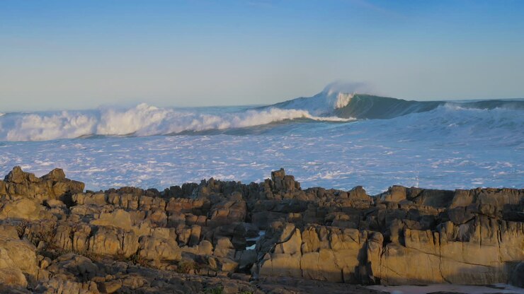 Onde enormi che si infrangono su una costa rocciosa, cielo azzurro.