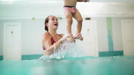 young mother is lifting her son from the water holding his feet while teaching him how to swim in the swimming pool. happy little boy and his mother are laughing and having fun