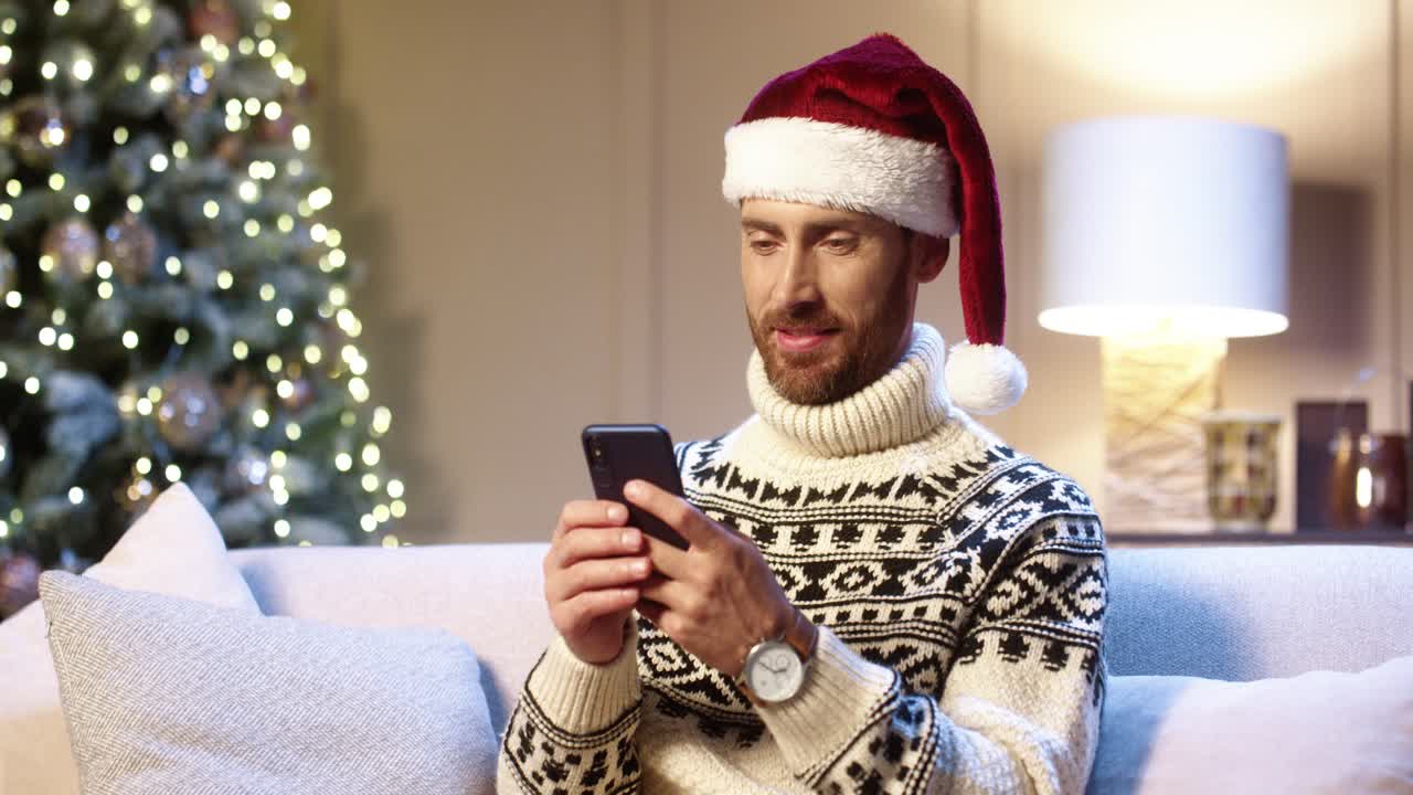 primer plano retrato de hombre guapo alegre con sombrero de santa sentado en una habitación acogedora cerca del árbol de navidad escribiendo en el teléfono inteligente con cara sorprendida ganar lotería de navidad