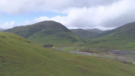 Aerial-reveal-shot-over-some-hills-to-a-stone-cottage-at-the-base-of-a-mountain-on-Rannoch-Moor-near-Loch-Treig