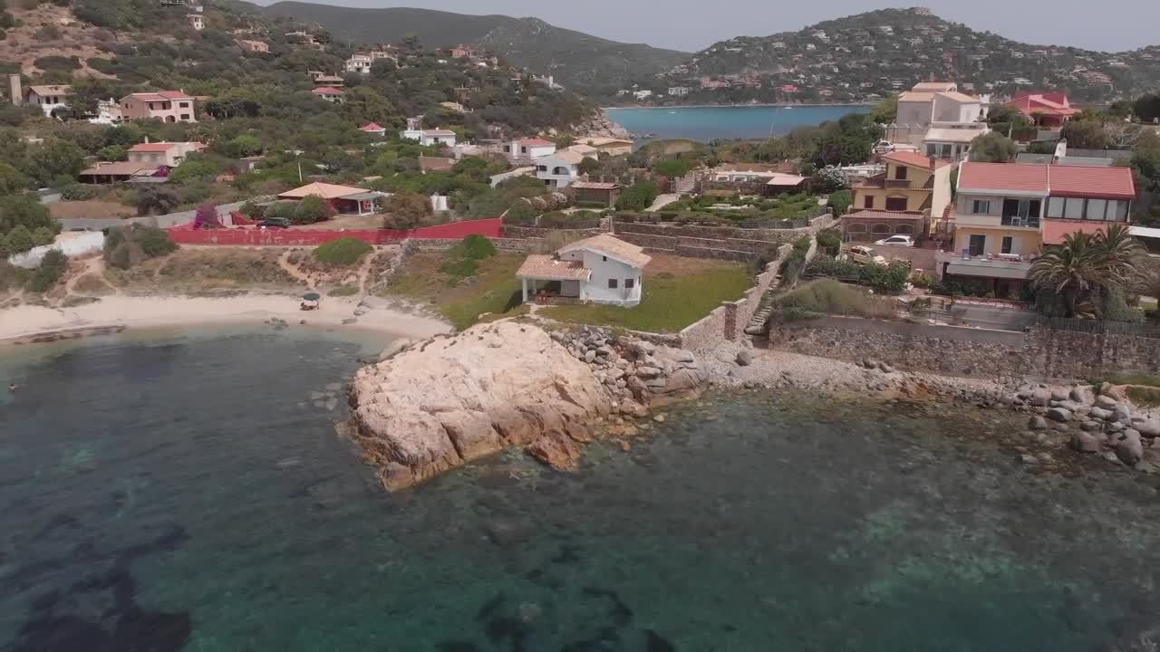Aerial drone view of coastal homes with red tiled roofs beside rocky shoreline and clear turquoise water in peaceful Mediterranean setting of Alghero Sardinia