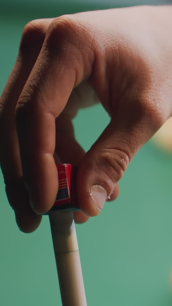 Close-up of person's hand chalking cue stick before taking precise shot on green pool table. Focus on preparation and technique with blurred billiard balls in background. Perfect for sports and gaming